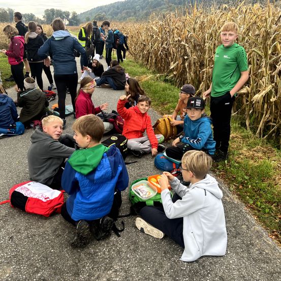 Mehrere junge Schüler sitzen am Rande eines Maisfeldwegs, einige stehen und reden. Ein Junge isst, während ein anderer Junge neben ihm steht.
