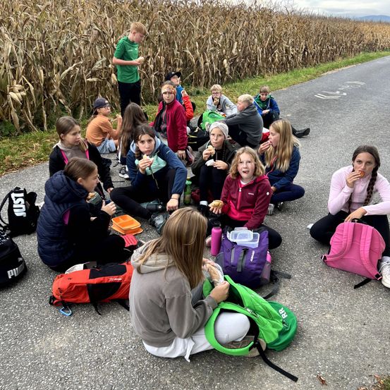 Eine Gruppe junger Menschen sitzt am Straßenrand und isst. Einige von ihnen tragen Jacken und Turnschuhe. Sie sind von Rucksäcken und Lunchboxen umgeben. In der Ferne steht ein Mann in der Nähe eines Maisfeldes.