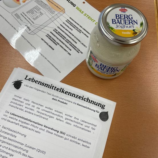 Ein Glas Vanille-Joghurt von Berg Bauern steht auf einem Holztisch mit zwei Informationsblättern. Ein Blatt hat deutschen Text über Nährwertinformationen, und das andere hat ein Etikett mit englischem Text.