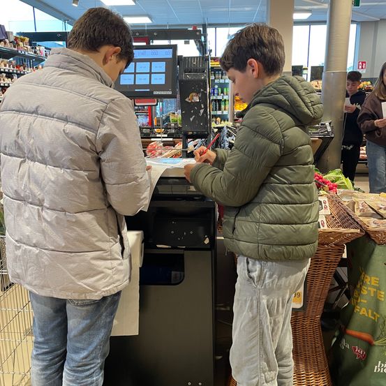 Zwei Jungen stehen an einer Kasse in einem Supermarkt. Ein Junge hält ein Papier, und der andere schaut auf einen Monitor.