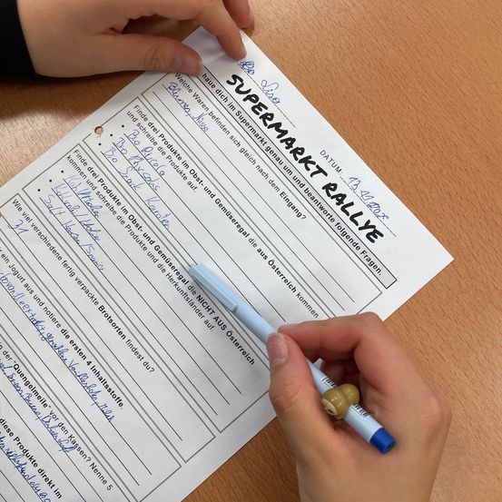 Eine Person füllt ein Formular mit der Überschrift 'SUPERMARKET RALLYE' mit einem Marker aus, schreibt auf ein Blatt Papier. Das Formular enthält Fragen und befindet sich auf einer Holzoberfläche.