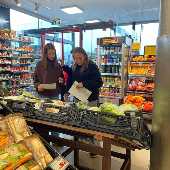 Zwei Frauen befinden sich in einem Supermarkt und betrachten ihre Einkaufslisten. Eine hält ein Papier in der Hand, und sie stehen in der Nähe eines Tisches mit frischem Gemüse, einschließlich Salat. Hinter ihnen sind Regale mit verschiedenen Lebensmitteln zu sehen.