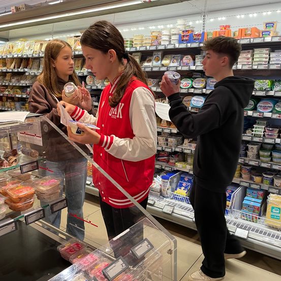 Drei Teenager sind in einem Lebensmittelgeschäft, eine Mädchen hält eine Dose und ein anderes Mädchen ein Papier, während der Junge einen Becher hält. Der Laden ist voll mit verschiedenen Produkten auf den Regalen.
