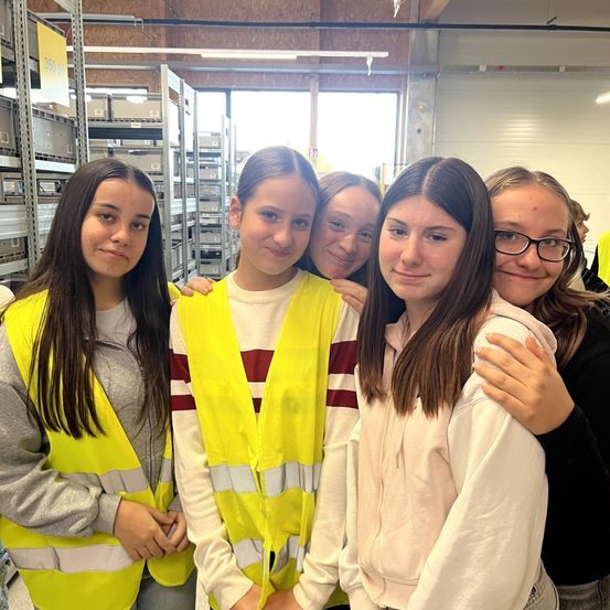 Fünf junge Frauen in gelben Sicherheitswesten stehen lächelnd und posieren für ein Foto in einem Lager. Dahinter befinden sich Regale mit Lagerboxen.