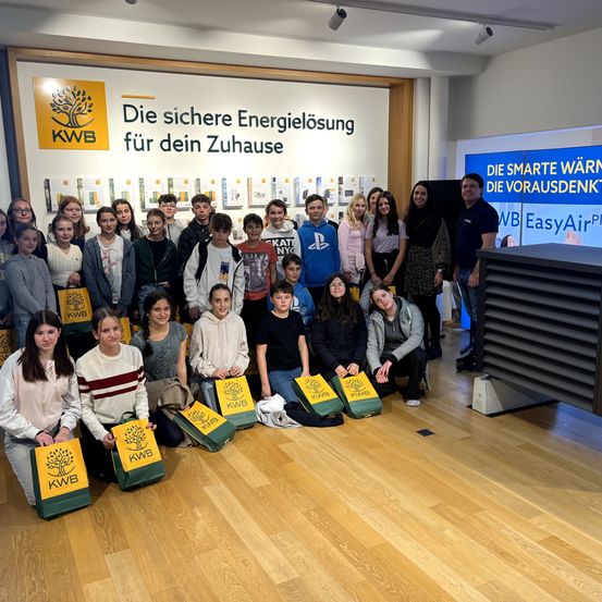 Eine Gruppe junger Menschen posiert mit KWB-Taschen vor einem Schild, in einem Gebäude mit Banner und Lampen.