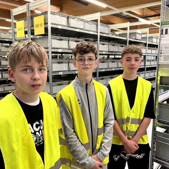 Drei Jungen in reflektierenden Westen stehen in einem Lager mit Regalen, die mit 360 01 und 350 12 beschriftet sind.
