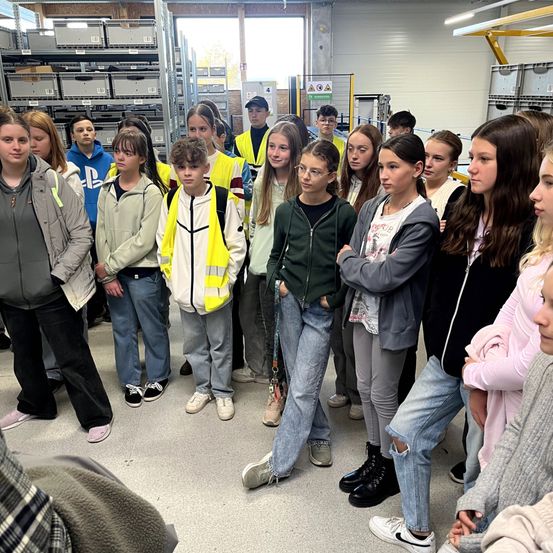 Eine Gruppe junger Menschen steht in einer Fabrik, einige tragen Sicherheitswesten. Sie stehen im Kreis und hören offenbar jemandem zu. Die Fabrik hat hohe Regale und einen hell beleuchteten Bereich.