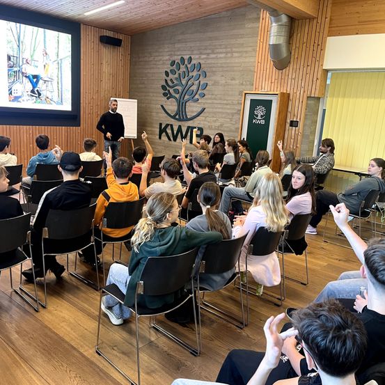 Eine Gruppe von Menschen sitzt in Stühlen in einem Raum mit Holzboden. Ein Mann steht vor ihnen an einer Tafel. Eine Projektionsleinwand zeigt ein Video. KWB-Markenzeichen sind an der Wand sichtbar.