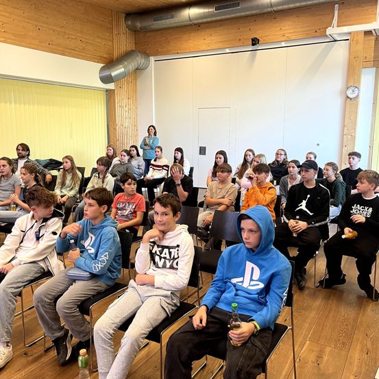 Eine Gruppe junger Menschen sitzt in Stühlen in einem Raum. Einige halten Getränke in der Hand, während andere sich unterhalten. Eine Frau steht in der Nähe der Wand.