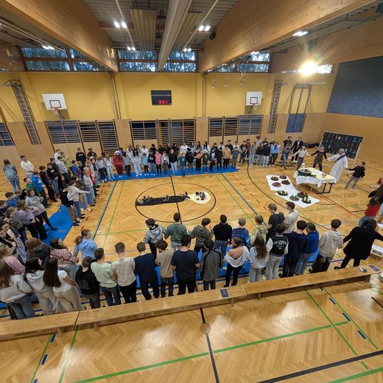 Eine große Gruppe von Menschen ist in einer Turnhalle versammelt. Einige stehen, andere sitzen auf Bänken. In der Mitte steht ein Tisch mit Essen.