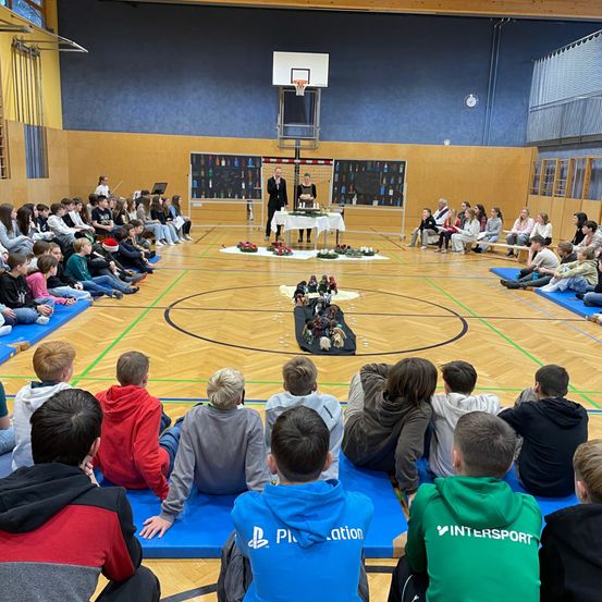 In einer Turnhalle versammeln sich eine Gruppe von Kindern und Erwachsenen um einen geschmückten Tisch in der Mitte und beobachten eine Aufführung. Zwei Personen stehen vor dem Tisch und führen ein Gespräch. Der Boden ist mit Matten ausgelegt, und die Turnhalle hat Holzböden und blaue Wände.