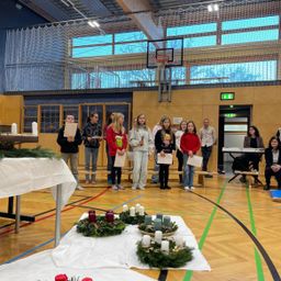 Eine Gruppe von Kindern steht in einer Basketballhalle mit Kränzen und Kerzen. Eine Frau sitzt auf einer Bank hinter ihnen, und Erwachsene stehen an den Seiten.