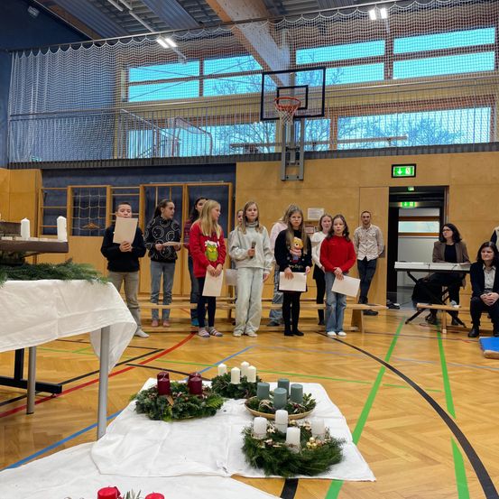 Eine Gruppe von Kindern steht in einer Basketballhalle mit Kränzen und Kerzen. Eine Frau sitzt auf einer Bank hinter ihnen, und Erwachsene stehen an den Seiten.