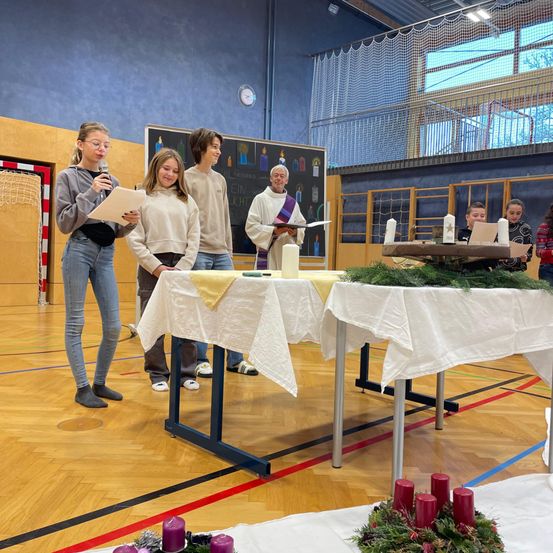 Eine Gruppe von Menschen steht um einen Tisch, der mit weißem Tuch und Grün bedeckt ist. Eine Person hält ein Mikrofon und Papier, während andere Kerzen halten. Sie befinden sich in einem Raum mit Holzböden und einer blauen Wand.
