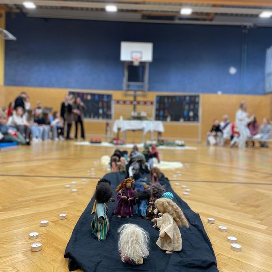 Indoor-Basketballplatz mit einer Weihnachtskrippe auf einem schwarzen Tuch. Kerzen umgeben die Szene, und Menschen sitzen an den Seiten.