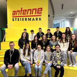 Eine Gruppe von Schülern sitzt auf einer gelben Plattform vor einem großen gelben Antennen Steiermark-Schild. Sie tragen alle Ausweise.