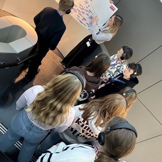 Eine Gruppe von Schülern in einem Wissenschaftslabor, die Kopfhörer tragen und einer Frau zuhören, die ein großes Diagramm hält.