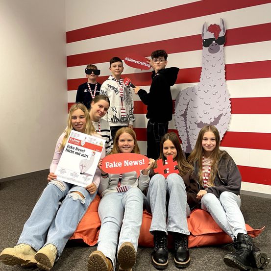 Eine Gruppe junger Menschen sitzt auf einem Sitzsack in einem Studio. Einer von ihnen hält ein Schild mit der Aufschrift Fake News. Hinter ihnen steht eine große weiß-rote Wand mit einer Ausschnitt eines lächelnden Alpakas.
