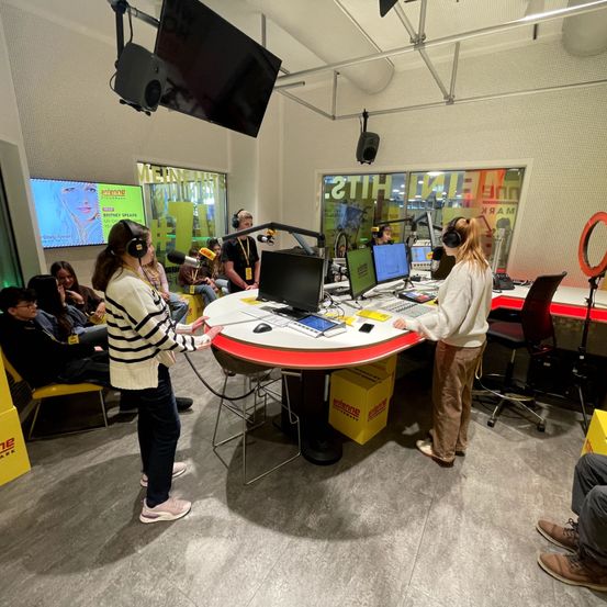 Zwei Frauen stehen hinter einem Schreibtisch in einem Studio, wahrscheinlich eine Aufnahme oder Sendung. Dahinter sitzen mehrere Personen in Stühlen und lauschen. Verschiedene Monitore, Mikrofone und Geräte sind auf dem Schreibtisch.