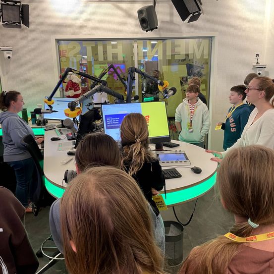 In einem Radiostudio versammeln sich mehrere Personen um einen Schreibtisch und beobachten einen Monitor. Eine Frau steht, während die anderen sitzen. Zwei Frauen und zwei Jungen sind anwesend.