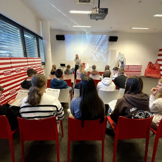 Eine Frau hält in einem Klassenzimmer eine Präsentation für eine Gruppe von Schülern, die auf roten Stühlen sitzen. Eine Projektionsleinwand zeigt ein Stadtgebäude, und der Raum hat weiße Wände und rot-weiß gestreifte Boxen.