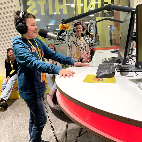 Ein Junge mit Kopfhörern spricht in ein Mikrofon in einem Studio mit Schreibtisch, Computer und anderen Personen.