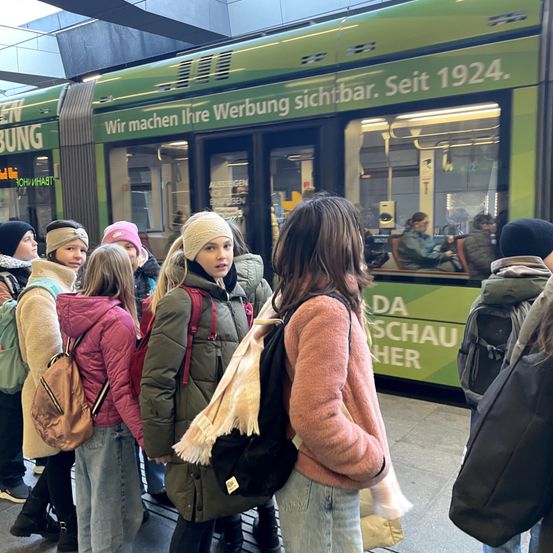 Eine Gruppe von Menschen, darunter junge Mädchen, steht in einer Schlange am Bahnhof und wartet auf eine grüne Straßenbahn. Die Straßenbahn hat Werbung an ihrer Seite.
