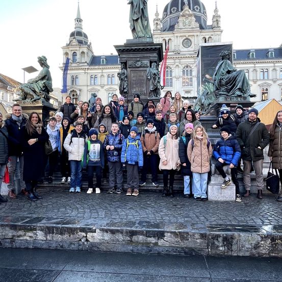 Eine Gruppe von Menschen posiert vor einer Statue auf einer Freiluft-Plaza, mit einem großen Gebäude und weiteren Statuen im Hintergrund.