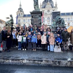 Eine Gruppe von Menschen posiert vor einer Statue auf einer Freiluft-Plaza, mit einem großen Gebäude und weiteren Statuen im Hintergrund.