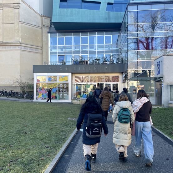 Mehrere Personen gehen auf einem Bürgersteig vor dem Next Liberty-Gebäude. Das Gebäude hat Glaswände und einen Balkon mit Pflanzen. Vor dem Gebäude geht eine Person in der Nähe geparkter Fahrräder. Der Bürgersteig ist von Gras umgeben.