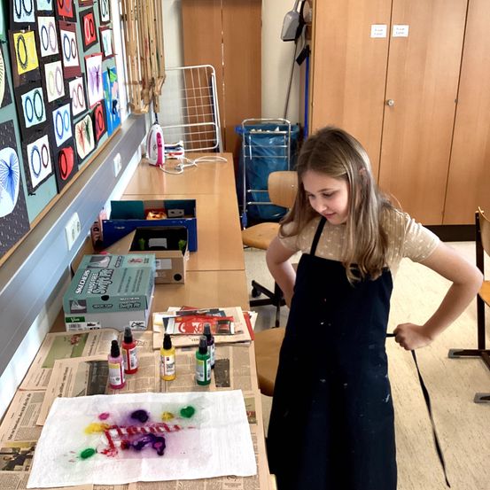 Ein junges Mädchen in einer Schürze steht vor einem Tisch mit Kunstmaterialien. Bunte Farbe liegt auf einem Papier auf dem Tisch. Hinter ihr zeigt eine Wand verschiedene Kunstwerke.