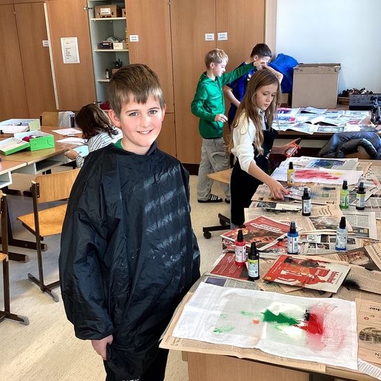 Ein Klassenzimmer mit Schülern, die an einem Malprojekt arbeiten. Ein junger Junge in einem schwarzen Mantel steht lächelnd, während andere Schüler auf Tischen malen, die mit Zeitungen bedeckt sind.