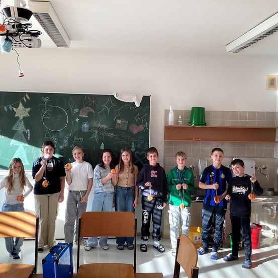 Eine Gruppe von Schülern steht in einem Klassenzimmer zusammen. Sie halten kleine Gegenstände, möglicherweise Auszeichnungen, in den Händen. Der Raum hat eine Tafel mit Zeichnungen, Stühle und ein Regal mit einem grünen Eimer.