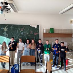 Eine Gruppe von Schülern steht in einem Klassenzimmer zusammen. Sie halten kleine Gegenstände, möglicherweise Auszeichnungen, in den Händen. Der Raum hat eine Tafel mit Zeichnungen, Stühle und ein Regal mit einem grünen Eimer.