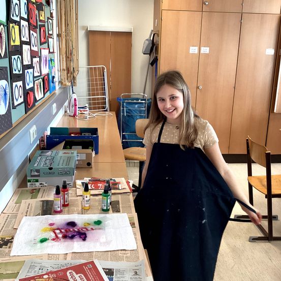 Ein junges Mädchen in einem Schürze steht in einem Klassenzimmer, lächelt und hat Kunstmaterial auf einem mit Zeitungen bedeckten Tisch. Kunstprojekte schmücken die Wand hinter ihr.