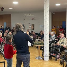 Ein älteres Paar spielt Musikinstrumente in einem Raum mit einer großen Menschenmenge älterer Menschen, die auf Stühlen und Rollstühlen sitzen. Der Raum ist mit Lichtern dekoriert.