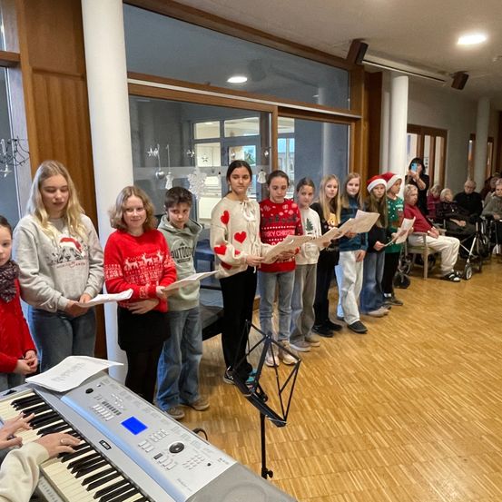 Eine Gruppe von Kindern in festlichen Pullovern singt mit Notenblättern in einem Raum mit Klavier, während ältere Erwachsene beobachten.