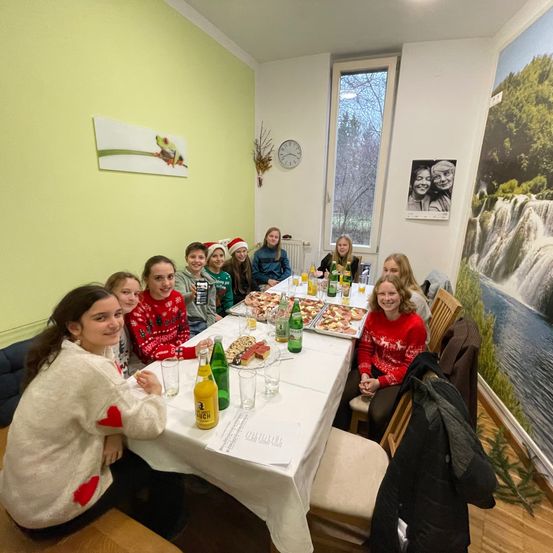Eine Gruppe von Frauen in Weihnachtspullovern sitzt um einen Tisch mit Pizza, Getränken und Gläsern. Der Tisch ist mit einem weißen Tuch bedeckt. Der Raum hat gelbe Wände, ein Fenster mit Blick auf einen Baum und zwei große Gemälde.