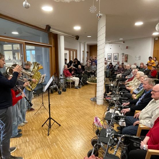 Eine Gruppe Musiker tritt auf einer Bühne für ein Publikum aus älteren Menschen in einem Raum mit Holzböden und weißen Wänden auf.