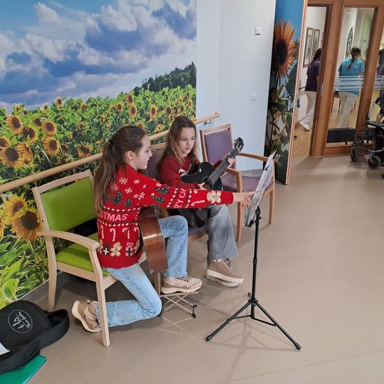 Zwei Mädchen spielen Gitarre. Ein Mädchen kniet und hält einen Notenständer, das andere sitzt. Sie sind in einem Raum mit einer Sonnenblumenwand und Stühlen. Im Hintergrund sind Menschen und ein Rollstuhl zu sehen.
