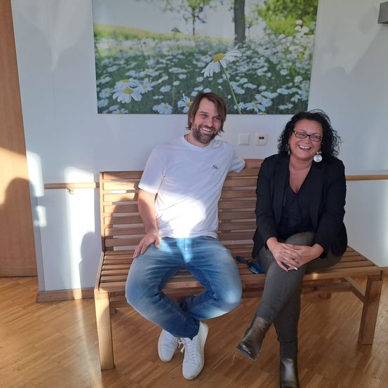 Ein Mann und eine Frau sitzen auf einer Bank in einem Raum und lächeln für ein Foto. Die Wand hinter ihnen zeigt ein großes Gemälde eines Feldes mit Gänseblümchen.