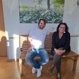 Ein Mann und eine Frau sitzen auf einer Bank in einem Raum und lächeln für ein Foto. Die Wand hinter ihnen zeigt ein großes Gemälde eines Feldes mit Gänseblümchen.