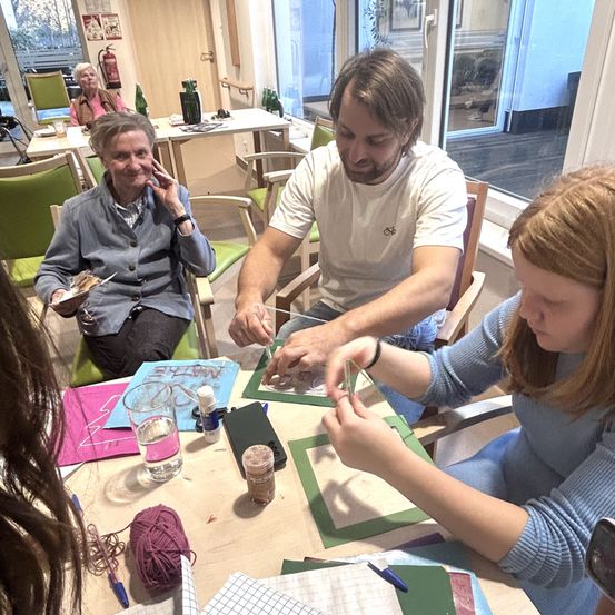 Eine Gruppe von Menschen bastelt an einem Tisch, darunter eine ältere Frau, ein Mann und ein Mädchen. Der Tisch ist mit Wolle, Scheren und einem Glas Wasser übersät. Eine weitere ältere Frau sitzt in der Nähe, und ein Feuerlöscher ist im Hintergrund zu sehen.