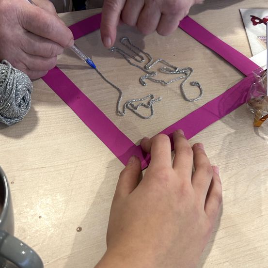 Ein Erwachsener und ein Kind erstellen ein Drahtkunstwerk auf einer rosa Unterlage auf einem Holztisch, mit einer Spule Garn und einem Becher in der Nähe.