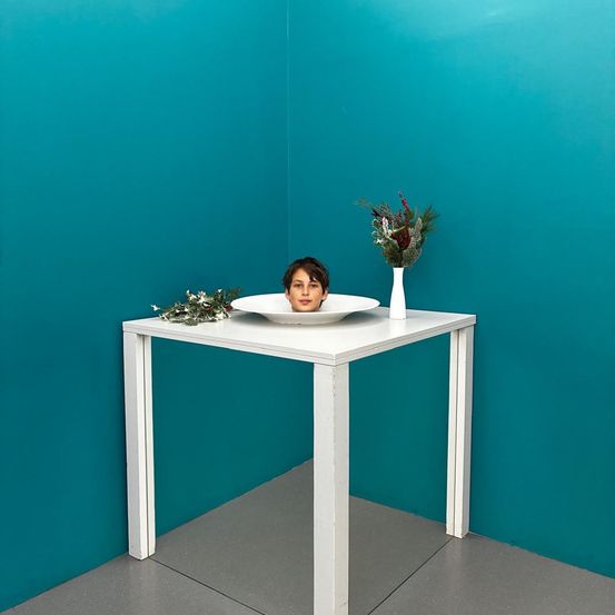 Ein junger Junge sitzt in einer weißen Schüssel auf einem Tisch mit Blumen in einer Vase an der Wand hinter ihm.