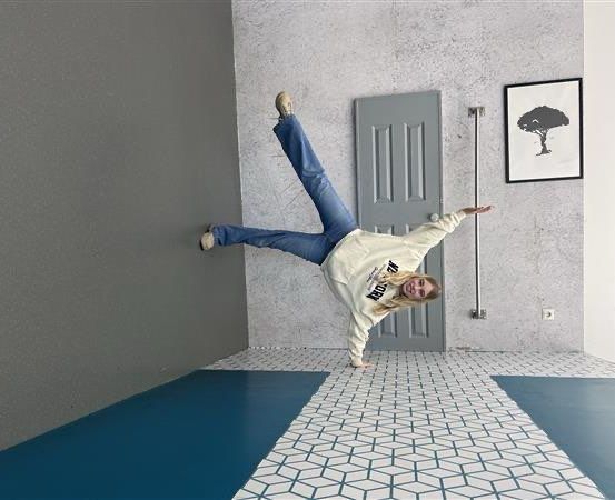 Eine Frau macht einen Handstand in einem Raum mit grauen Wänden, einem weißen Boden und einer Tür. Sie trägt ein weißes Sweatshirt und blaue Jeans.