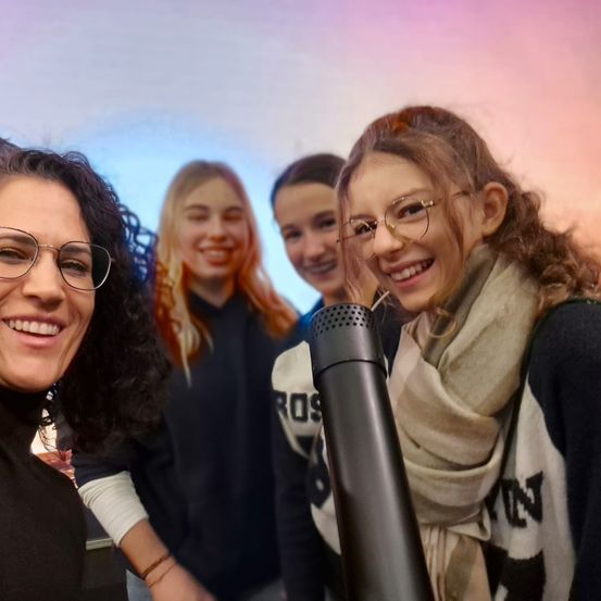 Eine Frau mit Brille und lockigem Haar lächelt neben einem Mikrofon. Hinter ihr lächeln drei Mädchen, eines von ihnen trägt einen Schal. Sie stehen vor einem bunten Hintergrund.