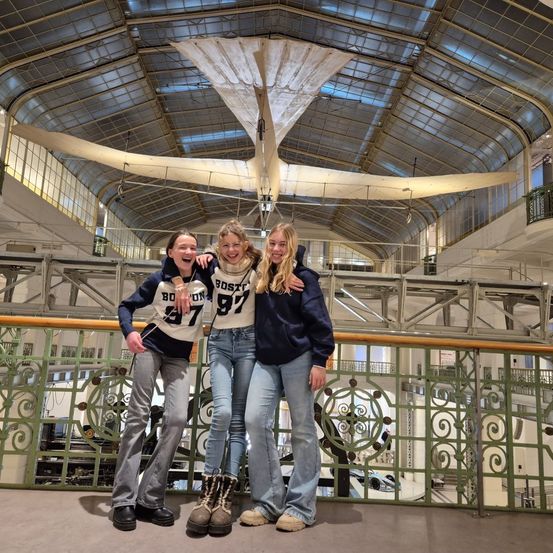 Drei Personen in einem Museum posieren unter einem Flugzeug mit der Nummer '97' auf ihren Shirts, lächeln und machen ein Foto.