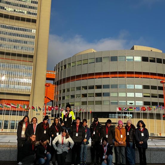 Eine Gruppe von Menschen posiert für ein Foto vor einem Gebäude mit Fahnen auf Stangen. Das Gebäude hat ein modernes Design mit Glasfenstern.