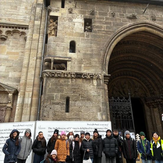 Eine Gruppe von Menschen in Winterkleidung steht vor einer großen steinernen Kirche mit einem gewölbten Eingang und kunstvollen Schnitzereien. Die Kirche scheint ein historisches oder religiöses Wahrzeichen zu sein.